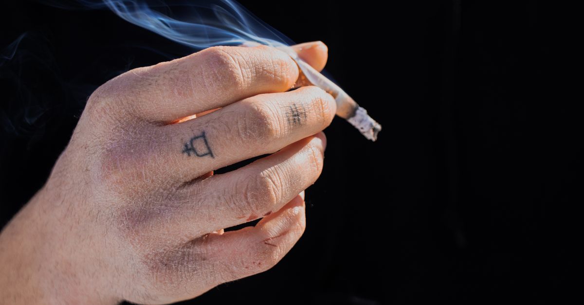 A detailed view of a tattooed hand holding a smoking cigarette, emphasizing the act of smoking.